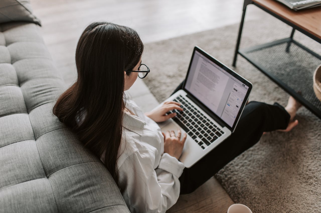 Woman working remotely with a laptop on the floor next to a sofa, enjoying comfortable home office setup.